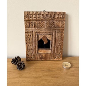 May include: A rectangular, intricately carved wooden wall mirror with a dark interior. The frame features detailed floral and geometric patterns. Two pinecones and a small candle sit on the wooden surface below the mirror.