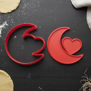 Pode incluir: Dois cortadores de biscoitos vermelhos em forma de lua crescente com um coração, junto com massa enrolada, farinha, ovos e um rolo em uma superfície preta. Imagem com tema de panificação.
