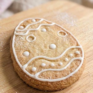 May include: An Easter egg-shaped biscuit decorated with white icing and dots, on a wooden board. The biscuit is light brown and dusted with icing sugar, creating a festive treat. The biscuit is approximately 10 cm long.