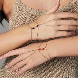 May include: Two hand chain bracelets. One bracelet is gold-toned with red beads, the other is silver-toned with black beads. The bracelets connect to the fingers with delicate chains. The background is a gold-colored, textured fabric.