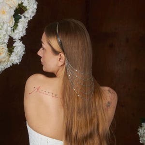 May include: A silver-toned metal hair accessory featuring a thin headband base with five delicate, cascading chains that drape over the side of the hair. The chains are composed of small, reflective circular links that create a layered effect.