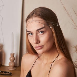 May include: A silver-toned headpiece featuring a delicate chain that rests across the forehead, accented with small, clear, teardrop-shaped crystals hanging at intervals along the chain, creating a subtle and elegant decorative hair accessory.