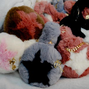 May include: A collection of fluffy, star-shaped keychains in various colors, including white, pink, gray, brown, and black. Some keychains feature gold-colored chain accents and charms. The keychains are made of soft, plush material.