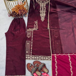 Puede incluir: Un conjunto de sat&eacute;n burdeos con una t&uacute;nica de manga larga con intrincados bordados dorados, pantalones a juego y una bufanda transparente. Sandalias marrones con correas decorativas y flores rosas completan el atuendo. La ropa se exhibe sobre una superficie gris claro.