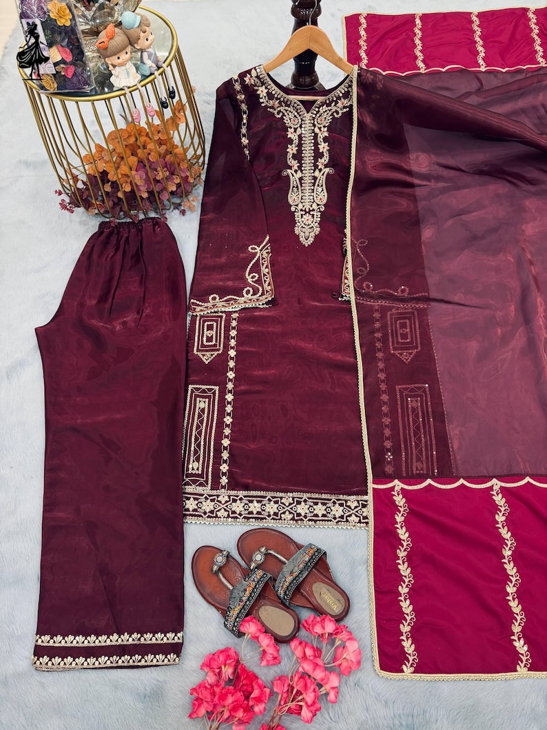 Puede incluir: Un conjunto de sat&eacute;n burdeos con una t&uacute;nica de manga larga con intrincados bordados dorados, pantalones a juego y una bufanda transparente. Sandalias marrones con correas decorativas y flores rosas completan el atuendo. La ropa se exhibe sobre una superficie gris claro.