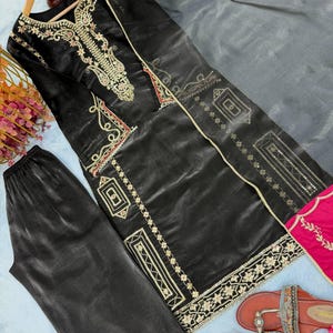 Puede incluir: Conjunto de kurta y pantal&oacute;n de sat&eacute;n negro con bordados dorados y detalles rosas. El kurta presenta intrincados detalles dorados alrededor del cuello y los bordes. Tambi&eacute;n se ven unas sandalias marrones con adornos.