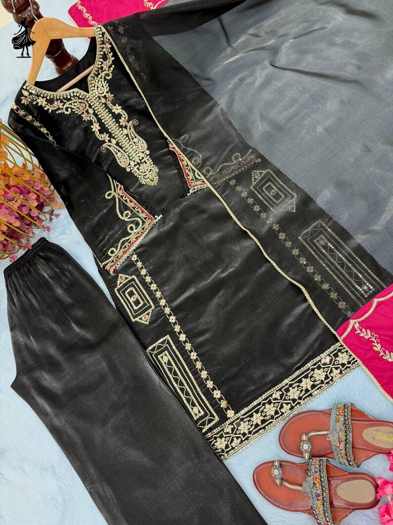 Puede incluir: Conjunto de kurta y pantal&oacute;n de sat&eacute;n negro con bordados dorados y detalles rosas. El kurta presenta intrincados detalles dorados alrededor del cuello y los bordes. Tambi&eacute;n se ven unas sandalias marrones con adornos.