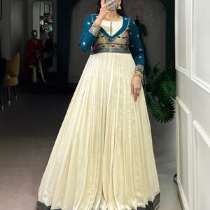 May include: A long, cream-colored dress with a pleated skirt and a teal bodice. The bodice has gold and silver accents, and the sleeves are long. The dress has a black and gold border at the hem.