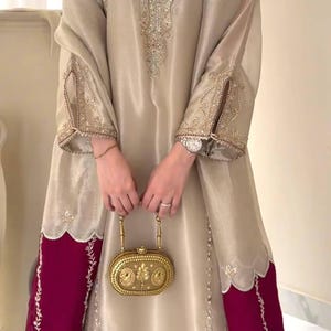 Puede incluir: Vestido de seda beige con intrincados bordados dorados en el corpi&ntilde;o y las mangas. El vestido presenta un dobladillo y pu&ntilde;os de manga de color rojo intenso en contraste. Se sostiene un clutch dorado en la parte delantera.