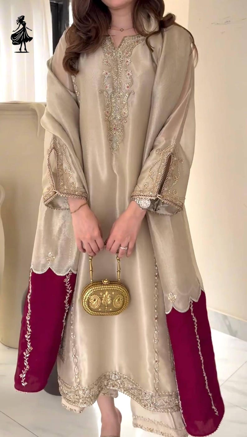 Puede incluir: Vestido de seda beige con intrincados bordados dorados en el corpi&ntilde;o y las mangas. El vestido presenta un dobladillo y pu&ntilde;os de manga de color rojo intenso en contraste. Se sostiene un clutch dorado en la parte delantera.