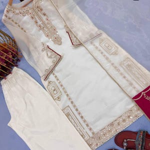 Puede incluir: Un conjunto de t&uacute;nica y pantal&oacute;n bordado en blanco roto. La t&uacute;nica presenta intrincados bordados dorados y rojos alrededor del cuello, los pu&ntilde;os y el dobladillo. El conjunto se combina con sandalias marrones. El conjunto se muestra sobre una superficie azul claro.
