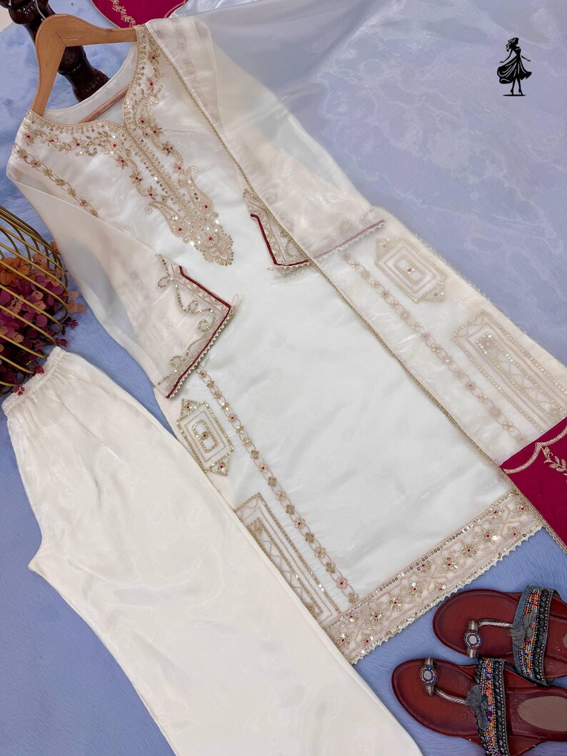 Puede incluir: Un conjunto de t&uacute;nica y pantal&oacute;n bordado en blanco roto. La t&uacute;nica presenta intrincados bordados dorados y rojos alrededor del cuello, los pu&ntilde;os y el dobladillo. El conjunto se combina con sandalias marrones. El conjunto se muestra sobre una superficie azul claro.