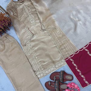 Puede incluir: Un conjunto beige bordado de tres piezas con pantal&oacute;n a juego y una bufanda transparente. El conjunto incluye una t&uacute;nica de manga larga con intrincados bordados plateados. Sandalias marrones con correas decorativas y flores rosas completan el conjunto.