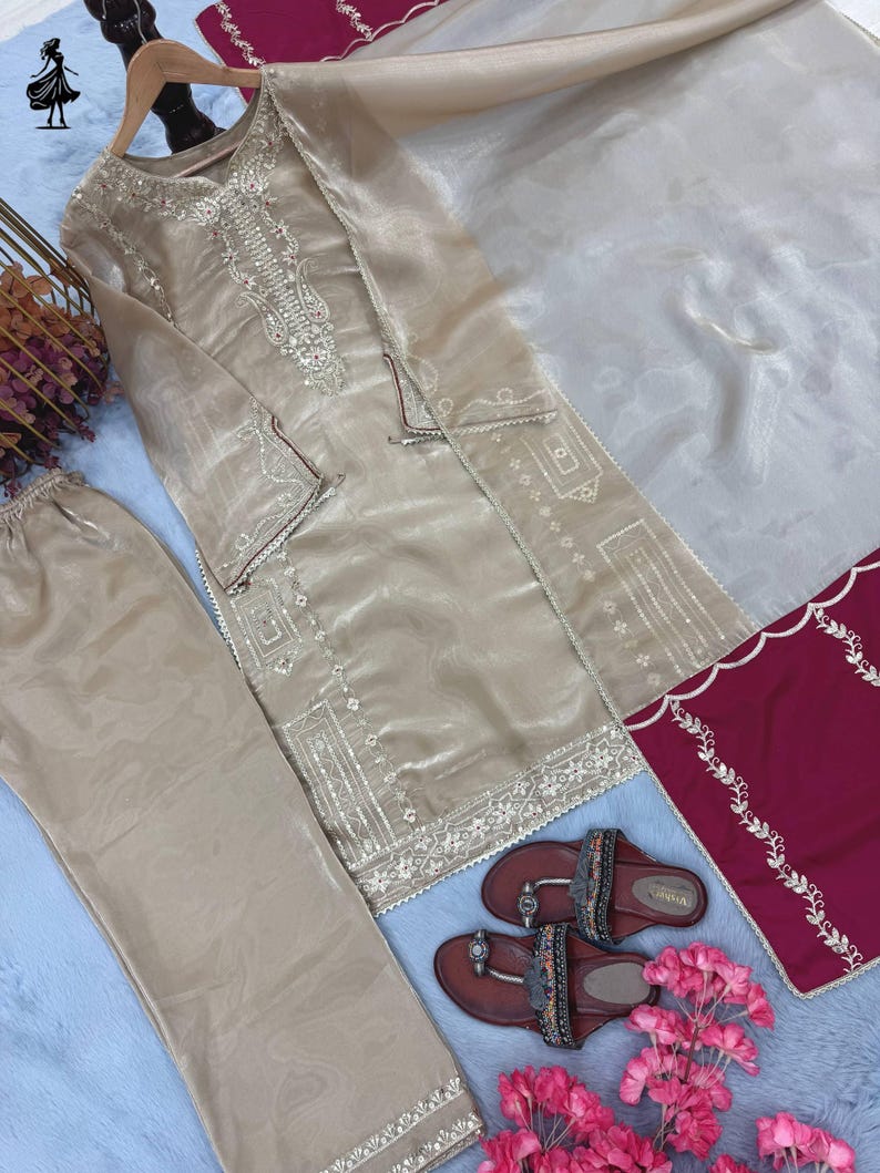 Puede incluir: Un conjunto beige bordado de tres piezas con pantal&oacute;n a juego y una bufanda transparente. El conjunto incluye una t&uacute;nica de manga larga con intrincados bordados plateados. Sandalias marrones con correas decorativas y flores rosas completan el conjunto.