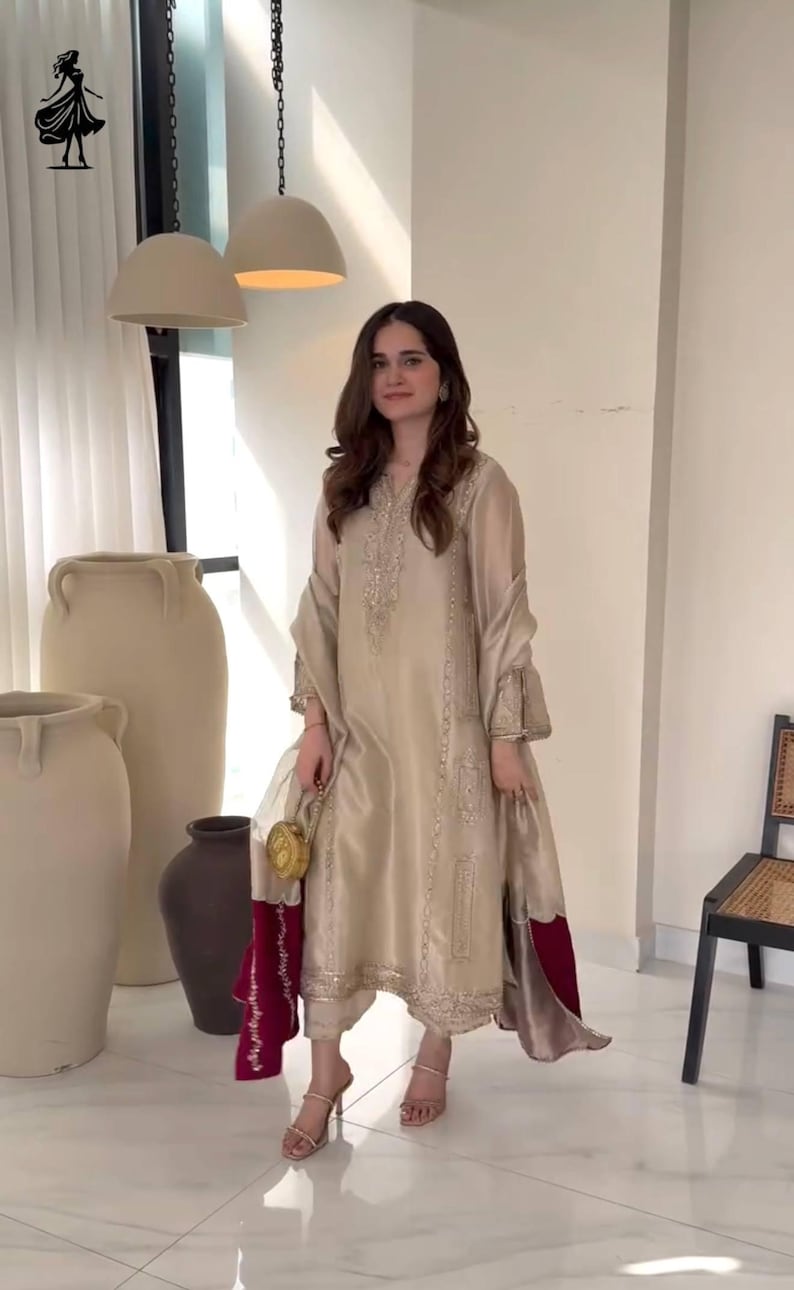 Puede incluir: Un conjunto salwar kameez de seda beige con bordados intrincados. El atuendo incluye una t&uacute;nica larga, pantalones a juego y una dupatta transparente. El conjunto se complementa con un peque&ntilde;o bolso dorado y sandalias de tac&oacute;n alto. Adecuado para ocasiones especiales.