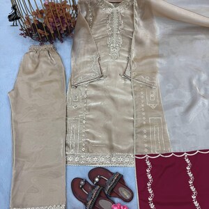 Puede incluir: Un conjunto beige bordado de dos piezas con pantal&oacute;n a juego y una blusa de manga larga. El conjunto incluye un chal transparente y se combina con sandalias marrones. El conjunto se exhibe sobre una superficie azul claro, con flores rosas y objetos decorativos.