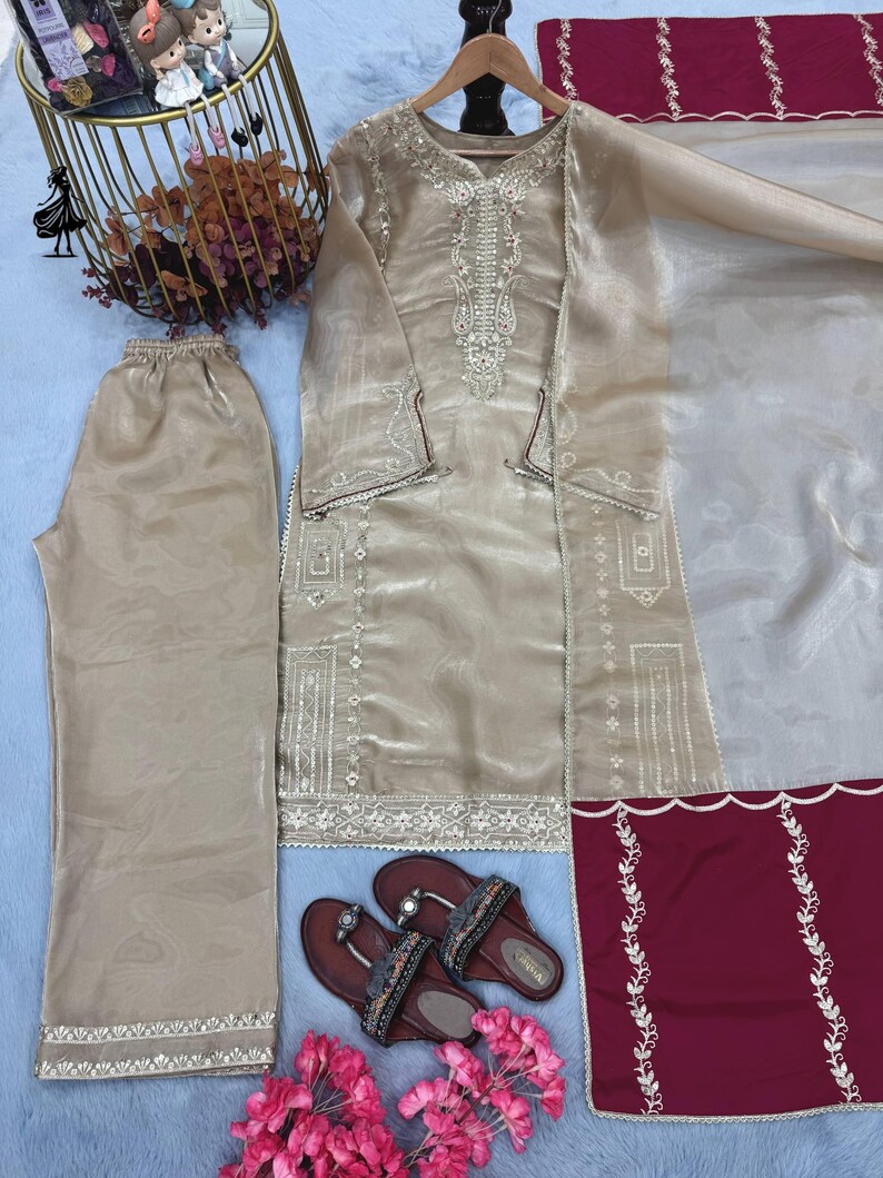Puede incluir: Un conjunto beige bordado de dos piezas con pantal&oacute;n a juego y una blusa de manga larga. El conjunto incluye un chal transparente y se combina con sandalias marrones. El conjunto se exhibe sobre una superficie azul claro, con flores rosas y objetos decorativos.