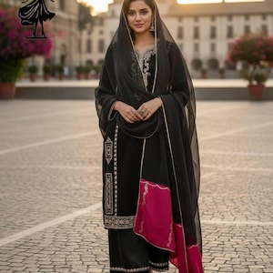 Puede incluir: Un conjunto de salwar kameez negro con bordados dorados y un borde rosa y dorado. El atuendo incluye un velo a juego. La ropa se usa al aire libre en un entorno urbano.