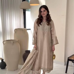 Puede incluir: Un traje kurta de seda beige con intrincados bordados dorados en el escote, las mangas y el dobladillo. El conjunto incluye pantalones a juego y una bufanda. La t&uacute;nica de manga larga se combina con sandalias doradas y un peque&ntilde;o bolso dorado.
