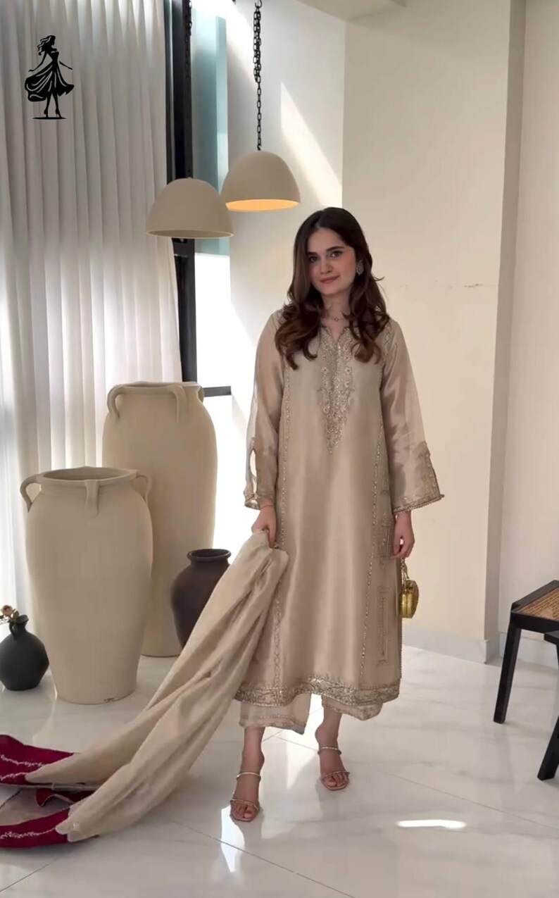 Puede incluir: Un traje kurta de seda beige con intrincados bordados dorados en el escote, las mangas y el dobladillo. El conjunto incluye pantalones a juego y una bufanda. La t&uacute;nica de manga larga se combina con sandalias doradas y un peque&ntilde;o bolso dorado.
