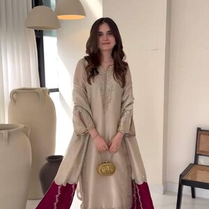 Puede incluir: Conjunto de seda beige con t&uacute;nica larga con bordados intrincados y mangas acampanadas. El atuendo incluye pantalones a juego y un chal con borde rojo oscuro. Un clutch dorado y sandalias completan el look. Adecuado para ocasiones especiales.