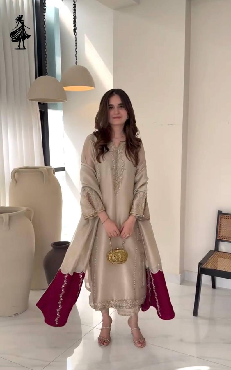 Puede incluir: Conjunto de seda beige con t&uacute;nica larga con bordados intrincados y mangas acampanadas. El atuendo incluye pantalones a juego y un chal con borde rojo oscuro. Un clutch dorado y sandalias completan el look. Adecuado para ocasiones especiales.