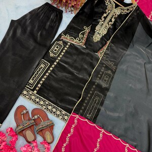 Puede incluir: Un conjunto bordado de terciopelo negro con pantal&oacute;n a juego, una bufanda transparente y una bufanda fucsia. El conjunto presenta bordados dorados y se combina con sandalias marrones. El conjunto est&aacute; dispuesto sobre una superficie azul claro con flores rosas.