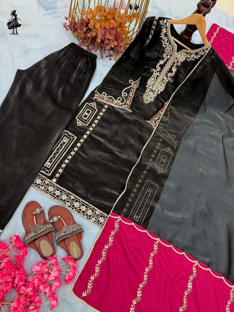 Puede incluir: Un conjunto bordado de terciopelo negro con pantal&oacute;n a juego, una bufanda transparente y una bufanda fucsia. El conjunto presenta bordados dorados y se combina con sandalias marrones. El conjunto est&aacute; dispuesto sobre una superficie azul claro con flores rosas.