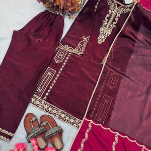 Puede incluir: Un conjunto de sat&eacute;n burdeos con una t&uacute;nica de manga larga con bordados dorados, pantalones a juego y una bufanda transparente. Sandalias marrones con correas decorativas y flores rosas completan el atuendo. La t&uacute;nica y los pantalones tienen ribetes dorados.
