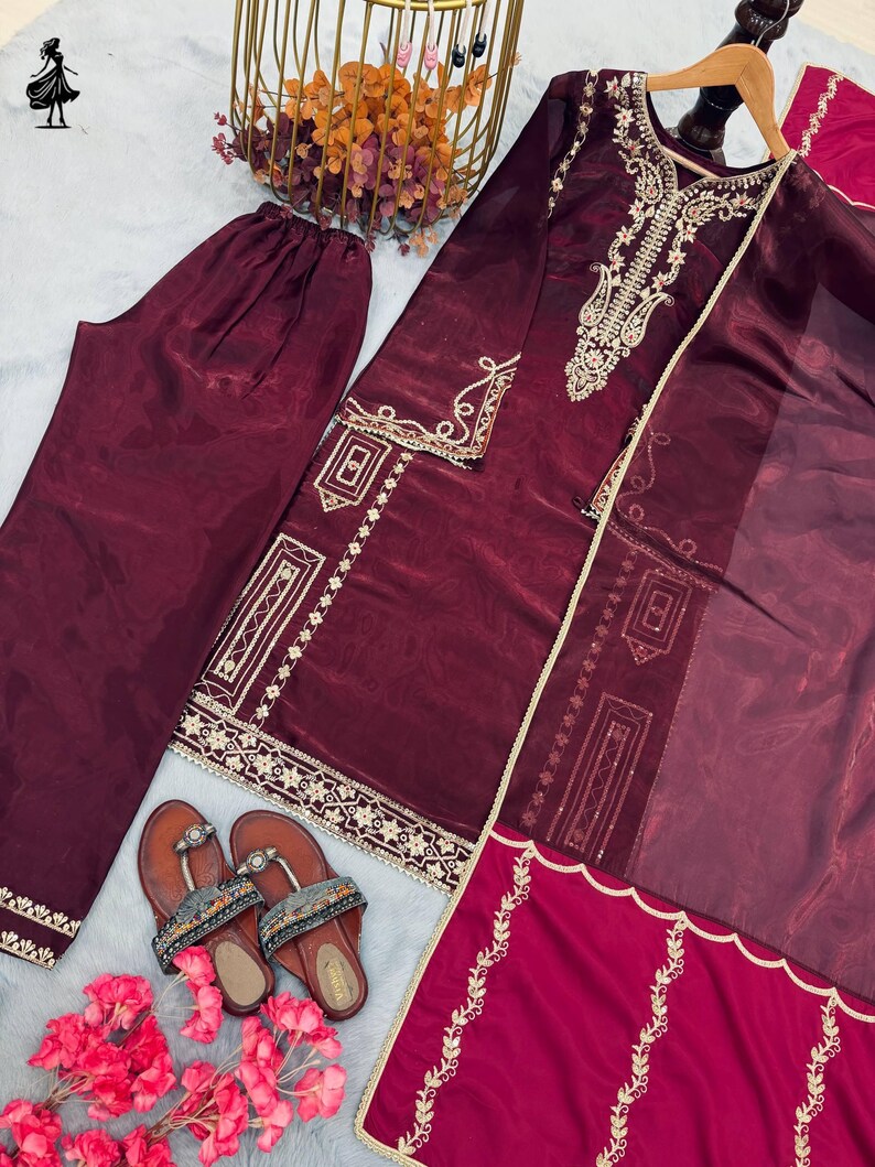Puede incluir: Un conjunto de sat&eacute;n burdeos con una t&uacute;nica de manga larga con bordados dorados, pantalones a juego y una bufanda transparente. Sandalias marrones con correas decorativas y flores rosas completan el atuendo. La t&uacute;nica y los pantalones tienen ribetes dorados.