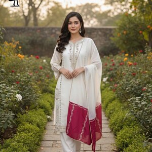 Puede incluir: Una mujer con un salwar kameez blanco bordado y un dupatta a juego. El dupatta tiene un borde granate. El atuendo se usa con sandalias. El fondo es un jard&iacute;n con un camino de piedra y plantas con flores.
