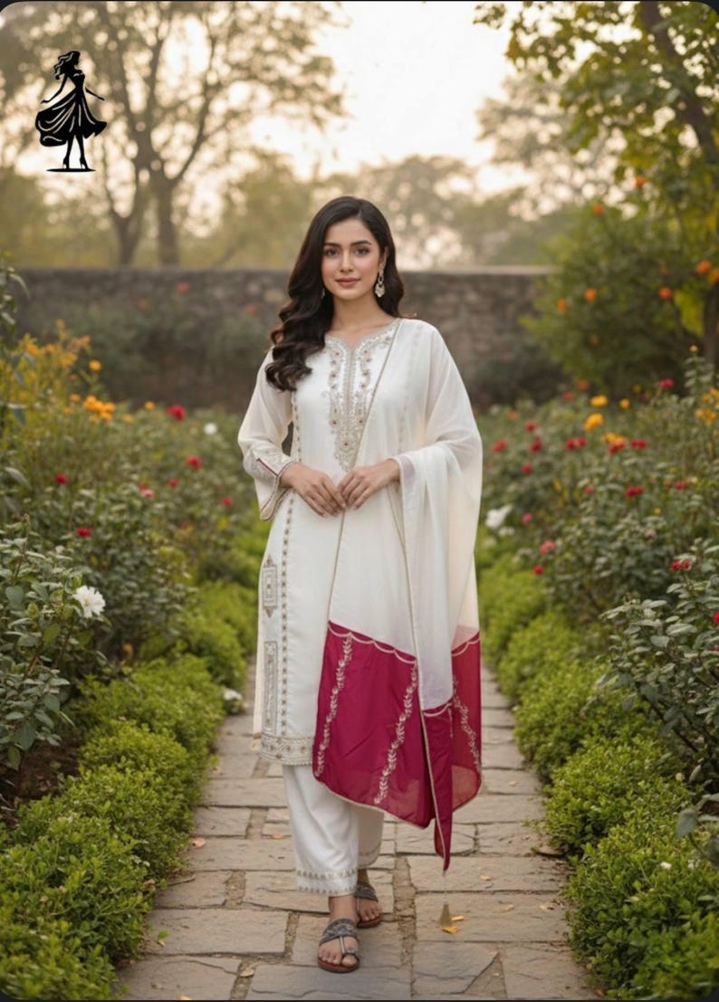 Puede incluir: Una mujer con un salwar kameez blanco bordado y un dupatta a juego. El dupatta tiene un borde granate. El atuendo se usa con sandalias. El fondo es un jard&iacute;n con un camino de piedra y plantas con flores.
