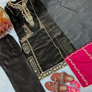 Puede incluir: Un conjunto de salwar kameez negro bordado con pantal&oacute;n a juego y una dupatta gris. El atuendo presenta bordados dorados y rojos. Sandalias marrones con correas decorativas y flores rosas completan el conjunto.