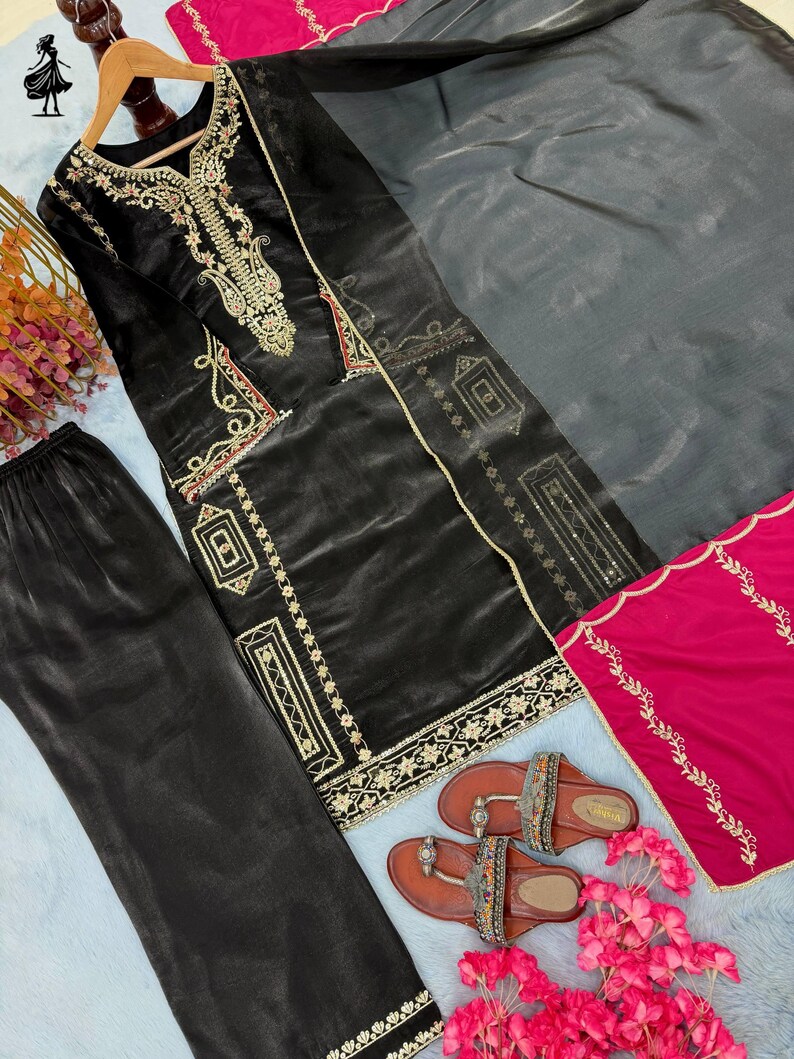 Puede incluir: Un conjunto de salwar kameez negro bordado con pantal&oacute;n a juego y una dupatta gris. El atuendo presenta bordados dorados y rojos. Sandalias marrones con correas decorativas y flores rosas completan el conjunto.