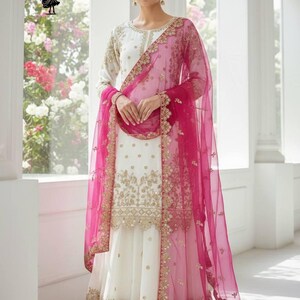 Peut inclure: Un ensemble salwar kameez blanc et or brodé avec un dupatta rose vif. La tenue présente des détails dorés complexes sur le corsage et l'ourlet, avec une écharpe en voile rose assortie.