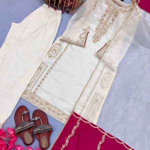 Puede incluir: Un conjunto de t&uacute;nica y pantal&oacute;n bordado en blanco roto con detalles dorados y granates. El conjunto incluye una bufanda granate con bordados dorados y sandalias marrones con adornos de cuentas. Tambi&eacute;n hay una cesta dorada con flores.