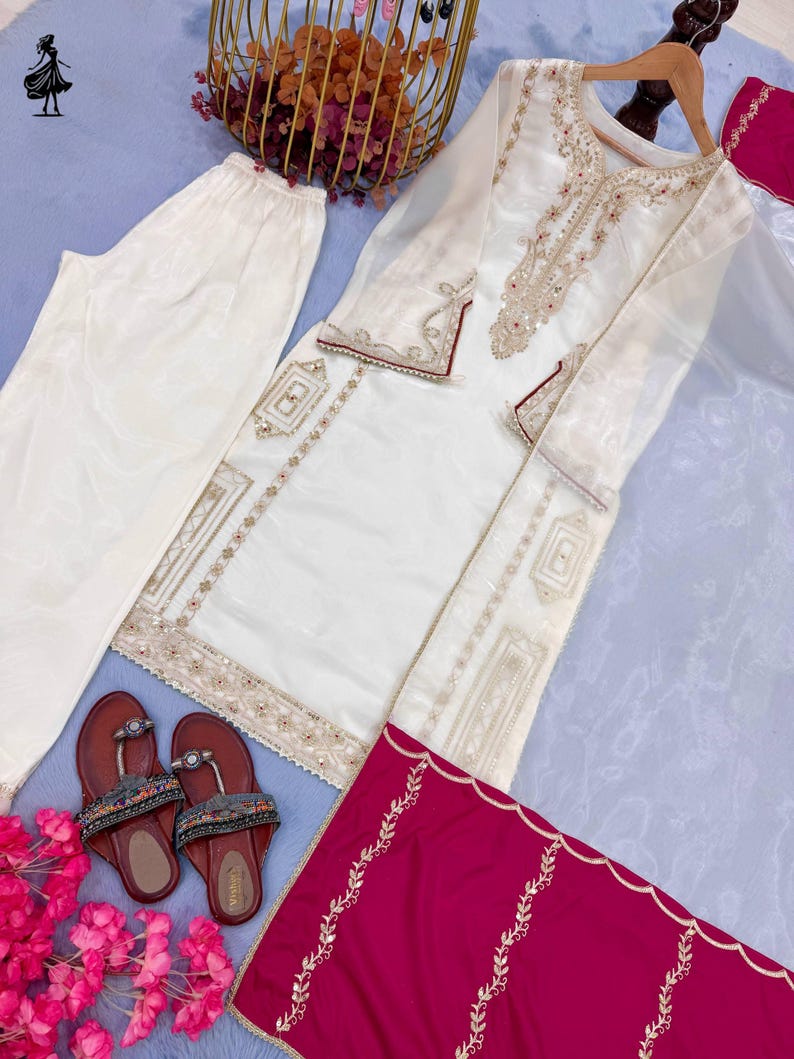 Puede incluir: Un conjunto de t&uacute;nica y pantal&oacute;n bordado en blanco roto con detalles dorados y granates. El conjunto incluye una bufanda granate con bordados dorados y sandalias marrones con adornos de cuentas. Tambi&eacute;n hay una cesta dorada con flores.