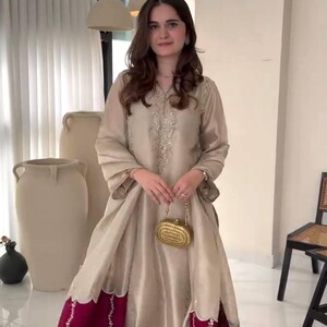 Puede incluir: Vestido de seda beige con detalles bordados y dise&ntilde;o fluido. El vestido presenta un dobladillo burdeos contrastante con intrincados bordados. Se sostiene un peque&ntilde;o bolso de mano dorado. El atuendo es adecuado para ocasiones especiales.