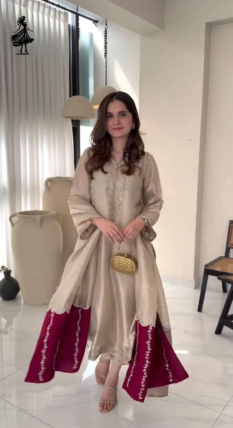 Puede incluir: Vestido de seda beige con detalles bordados y dise&ntilde;o fluido. El vestido presenta un dobladillo burdeos contrastante con intrincados bordados. Se sostiene un peque&ntilde;o bolso de mano dorado. El atuendo es adecuado para ocasiones especiales.
