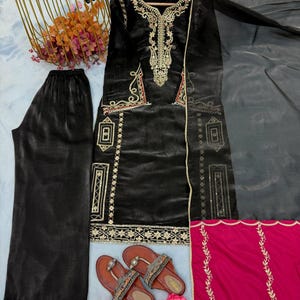 Puede incluir: Conjunto de t&uacute;nica y pantal&oacute;n negro de terciopelo bordado con detalles dorados y rojos. Incluye una bufanda gris transparente y una pieza de acento rosa brillante. Sandalias marrones con correas decorativas.