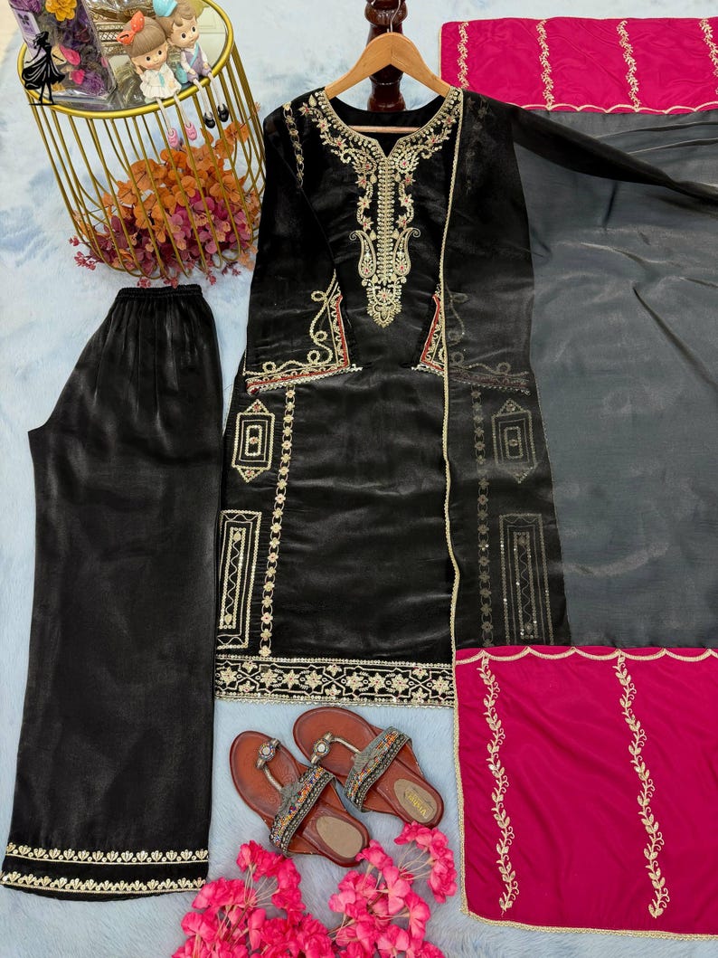 Puede incluir: Conjunto de t&uacute;nica y pantal&oacute;n negro de terciopelo bordado con detalles dorados y rojos. Incluye una bufanda gris transparente y una pieza de acento rosa brillante. Sandalias marrones con correas decorativas.