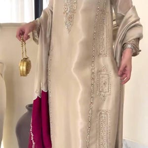 Puede incluir: Conjunto de seda beige con intrincados bordados plateados. El atuendo incluye una t&uacute;nica larga con detalles adornados, pantalones a juego y un dobladillo magenta en contraste. Un clutch dorado a&ntilde;ade un toque de elegancia.