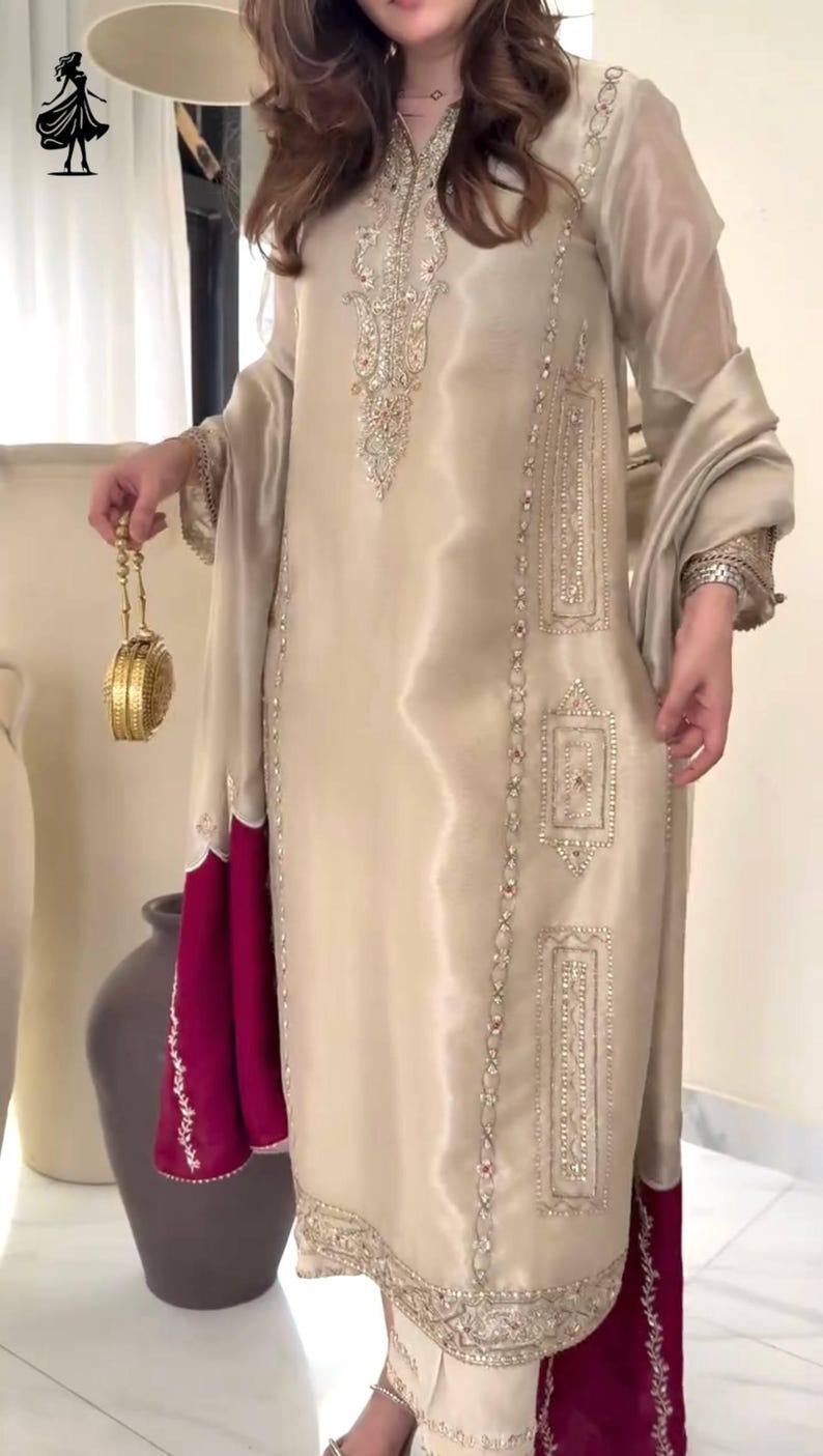 Puede incluir: Conjunto de seda beige con intrincados bordados plateados. El atuendo incluye una t&uacute;nica larga con detalles adornados, pantalones a juego y un dobladillo magenta en contraste. Un clutch dorado a&ntilde;ade un toque de elegancia.