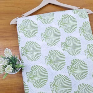 May include: A white fabric textile featuring a repeating pattern of light green scallop shells. The fabric is draped over a light-colored wooden clothes hanger, displaying a consistent print of sea-themed motifs across the entire surface area.
