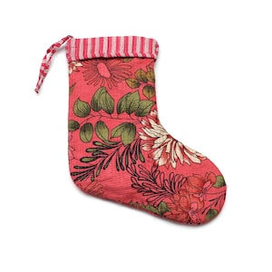 Puede incluir: Un calcetín navideño rojo con un estampado floral con hojas verdes y flores blancas y rojas. La parte superior del calcetín tiene rayas rojas y blancas, y un lazo a juego para colgar. El calcetín es de tela.