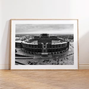 May include: Monochrome framed print of Lambeau Field, a large stadium with a circular design. The stadium's name is visible on the scoreboard. The image is set against a cloudy sky, with cars parked around the stadium.