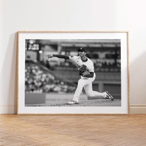 May include: Black and white framed print of a baseball pitcher in action, throwing a ball. The pitcher is wearing a baseball uniform and cap. The background shows a stadium filled with spectators.