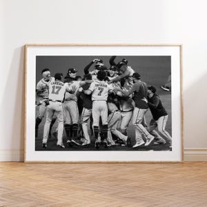Könnte beinhalten: Schwarz-Weiß-Druck in einem Rahmen, der ein Baseballteam beim Feiern zeigt. Spieler in Uniformen mit Nummern und Namen umarmen sich. Das Bild ist vor einem dunklen Hintergrund aufgenommen und fängt einen Moment des Triumphs ein.