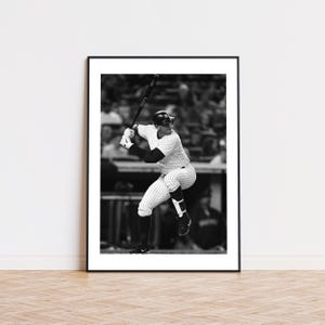 May include: Black and white framed print of a baseball player in a pinstripe uniform, poised to swing a bat. The player wears a helmet and batting gloves, captured mid-swing. The image is set against a blurred background of a stadium.
