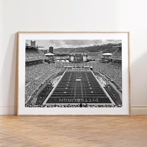 May include: Black and white framed print of a football stadium. The field is marked with yard lines and the word "PITTSBURGH". The stands are filled with spectators. A bridge and city skyline are visible in the background.